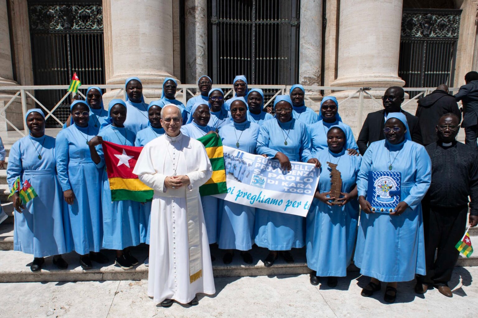 Les Sœurs de la Providence de Saint Paul de Kara à Rome avec le drapeau du Togo et la bannière « Suore della Provvidenza S. Paolo di Kara – Togo », aux côtés d’un ecclésiastique en soutane blanche