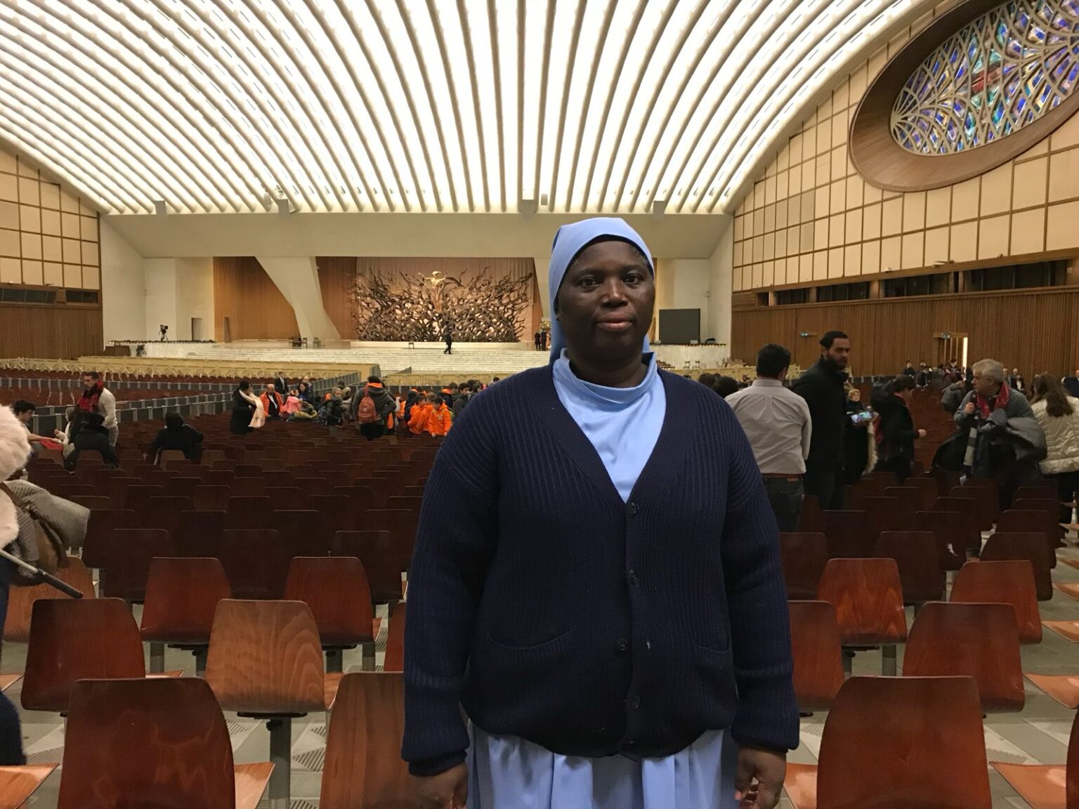 Une sœur en voile bleu et gilet sombre, debout dans une grande salle d’audience